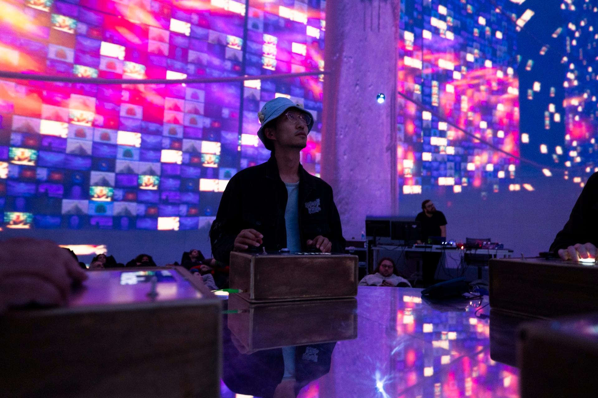 An audience member at a controller in front of a wall of glowing tiles